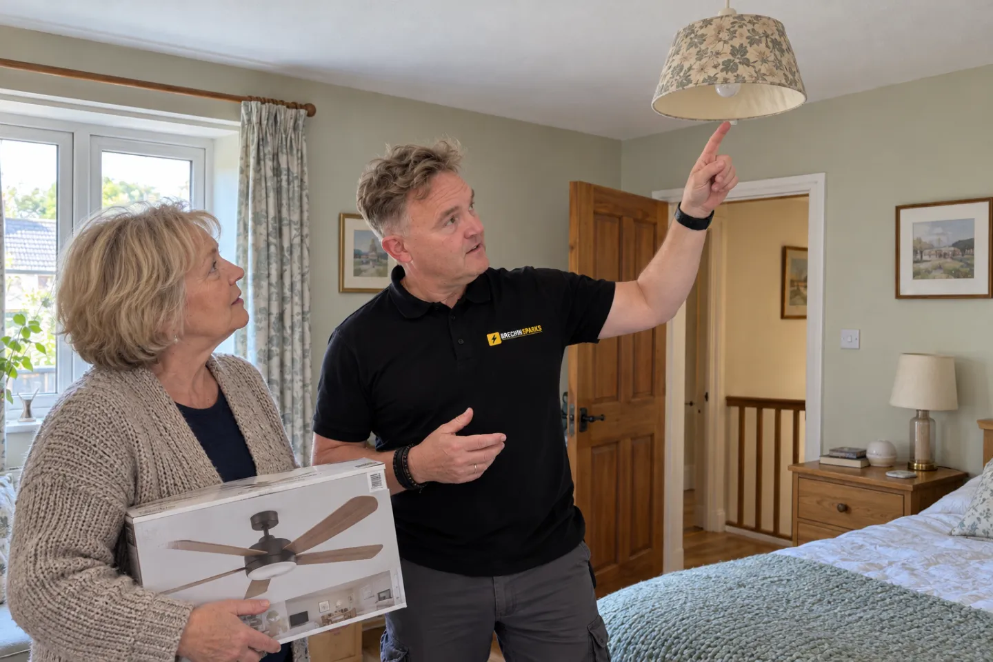 James and a homeowner holding a boxed fan, looking up at an old bedroom pendant